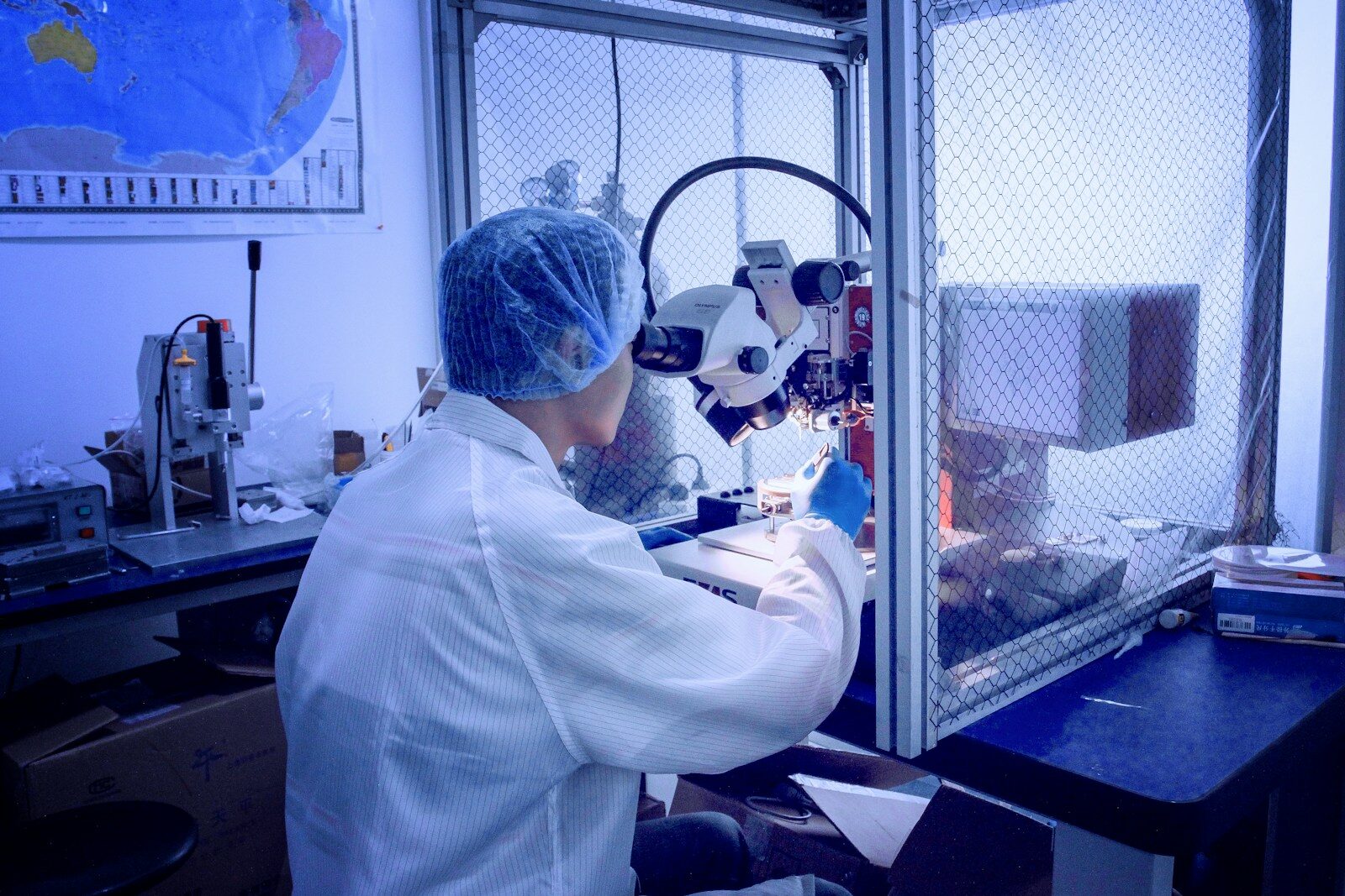 A researcher in a lab coat and hairnet examines samples under a microscope, illustrating a focused scientific analysis in a controlled environment.