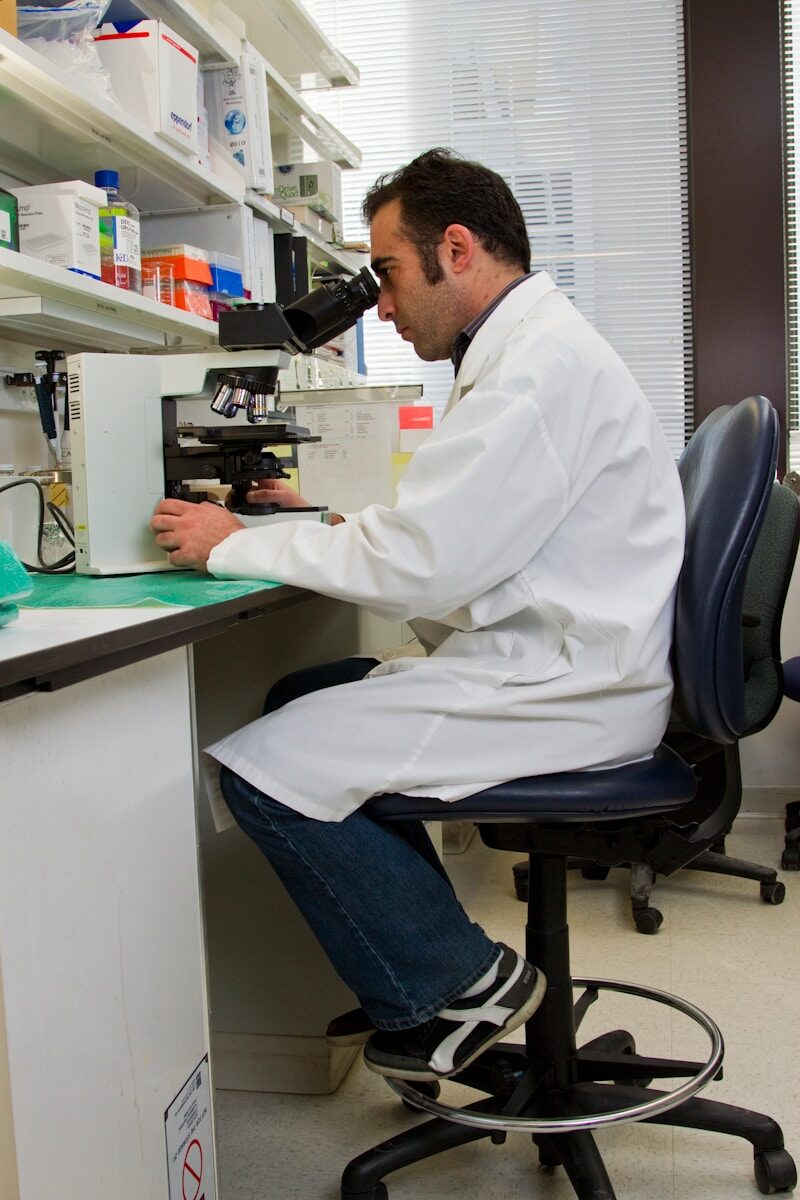 Scientist in a lab coat examines a sample under a microscope at a laboratory bench, surrounded by laboratory equipment and supplies.