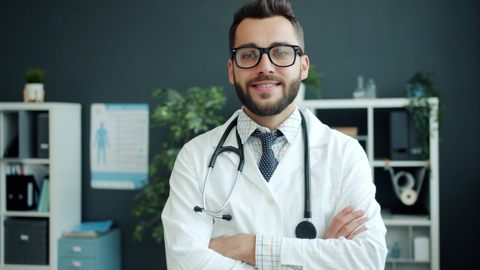 Smiling male doctor in a white coat and stethoscope stands confidently in a modern clinic, surrounded by shelves and plants.